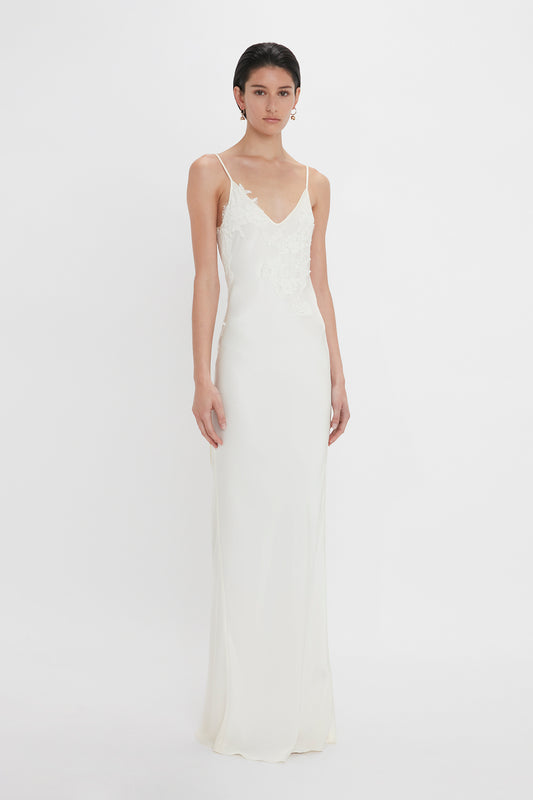 A woman stands against a white background, wearing an elegant sleeveless Exclusive Lace Detail Floor-Length Cami Dress in Ivory by Victoria Beckham.
