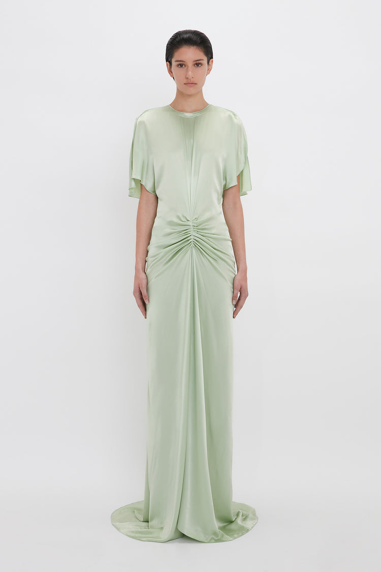A woman in a Victoria Beckham exclusive floor-length gathered dress in jade, with short sleeves and a ruched front, standing against a white background.