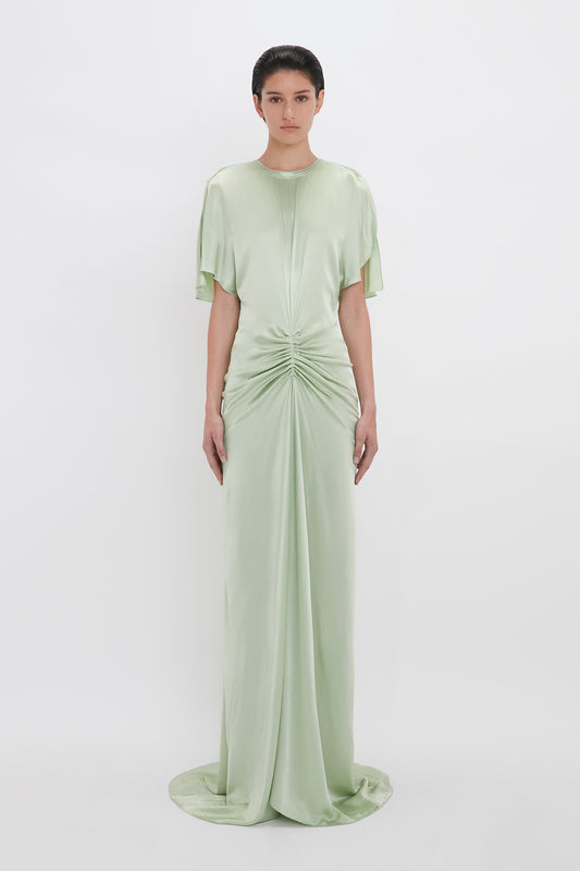 A woman in a Victoria Beckham exclusive floor-length gathered dress in jade, with short sleeves and a ruched front, standing against a white background.