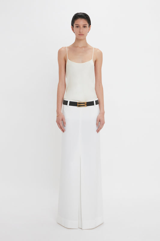 A person stands against a plain white background, wearing an ivory sleeveless top, an Exclusive Tailored Floor-Length Pleated Skirt In Ivory by Victoria Beckham, and a black belt with a gold buckle.