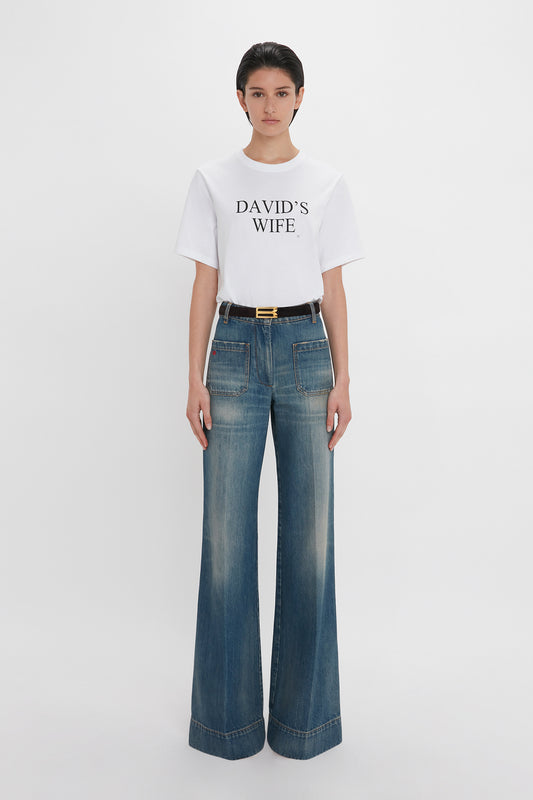 A woman wearing a white Victoria Beckham 'David's Wife' Slogan T-Shirt with "David's Wife" text and blue high-waisted, wide-leg jeans, paired with a black belt.