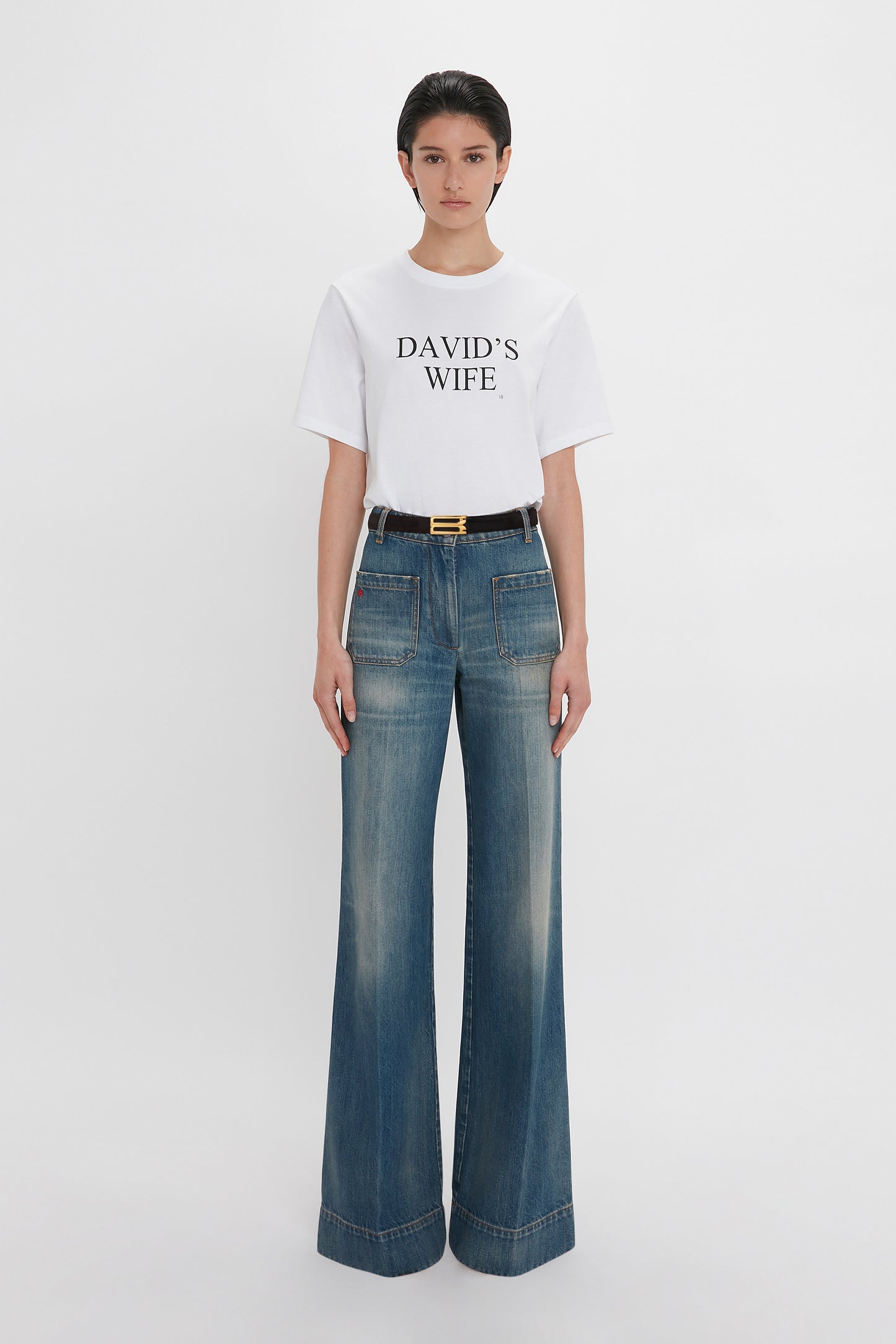 A woman wearing a white t-shirt with "david's wife" text and high-waisted Alina Jean In Indigrey Wash jeans by Victoria Beckham, accessorized with a black belt with a gold buckle, standing against a