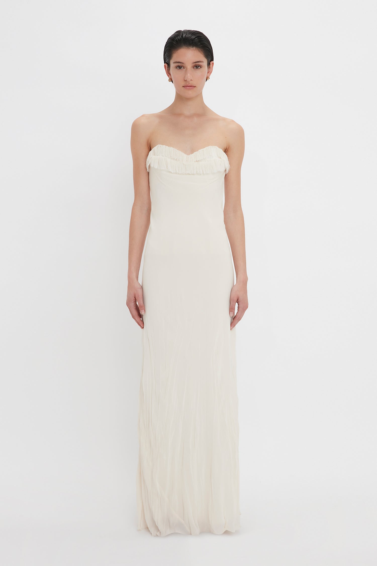 A woman stands facing the camera, wearing an elegant strapless cream gown with a softly pleated bust and a long, flowing crease-effect skirt by Victoria Beckham.