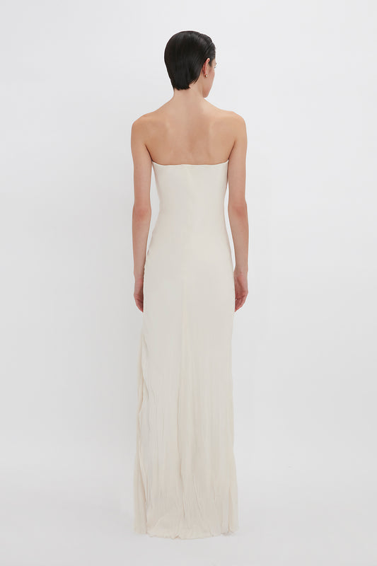 A woman with short dark hair stands with her back to the camera, wearing a Victoria Beckham Exclusive Floor-Length Corset Detail Gown in Ivory.