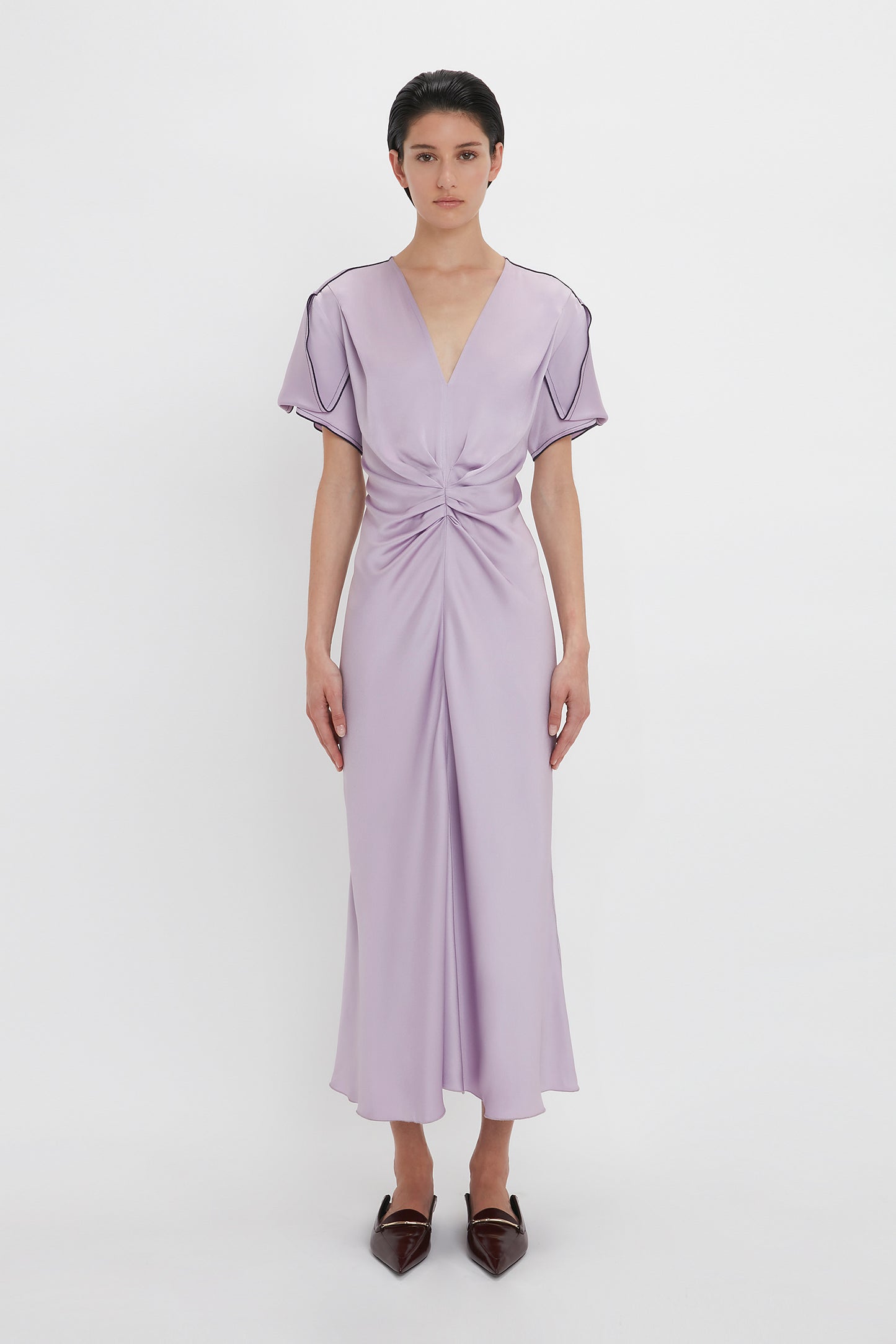 A person stands facing forward wearing the Gathered V-Neck Midi Dress In Petunia by Victoria Beckham, with a V-neck, short sleeves, and knotted waist detail. They are also wearing pointed-toe brown shoes. The background is plain white.