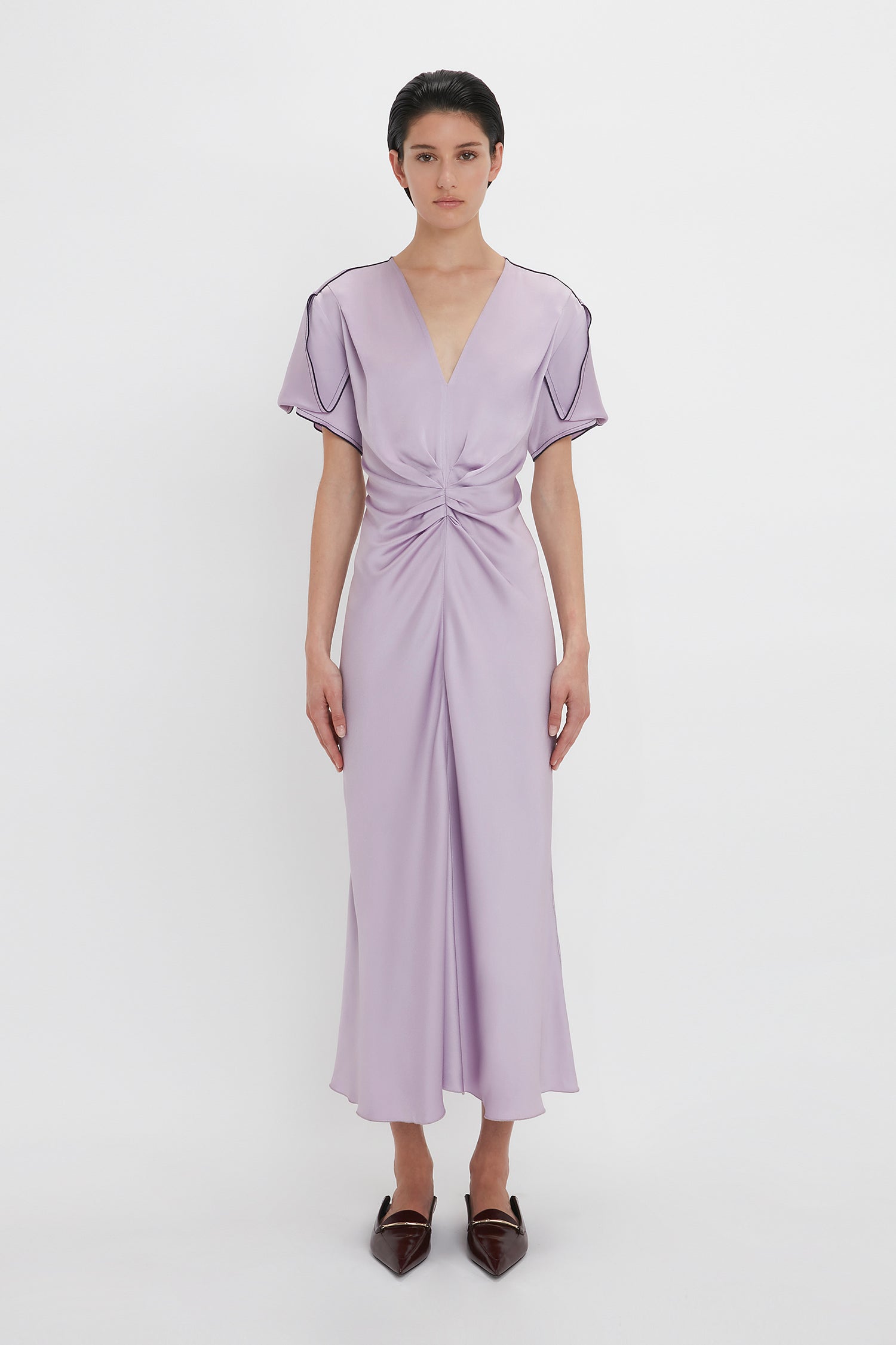 A person stands facing forward wearing the Gathered V-Neck Midi Dress In Petunia by Victoria Beckham, with a V-neck, short sleeves, and knotted waist detail. They are also wearing pointed-toe brown shoes. The background is plain white.