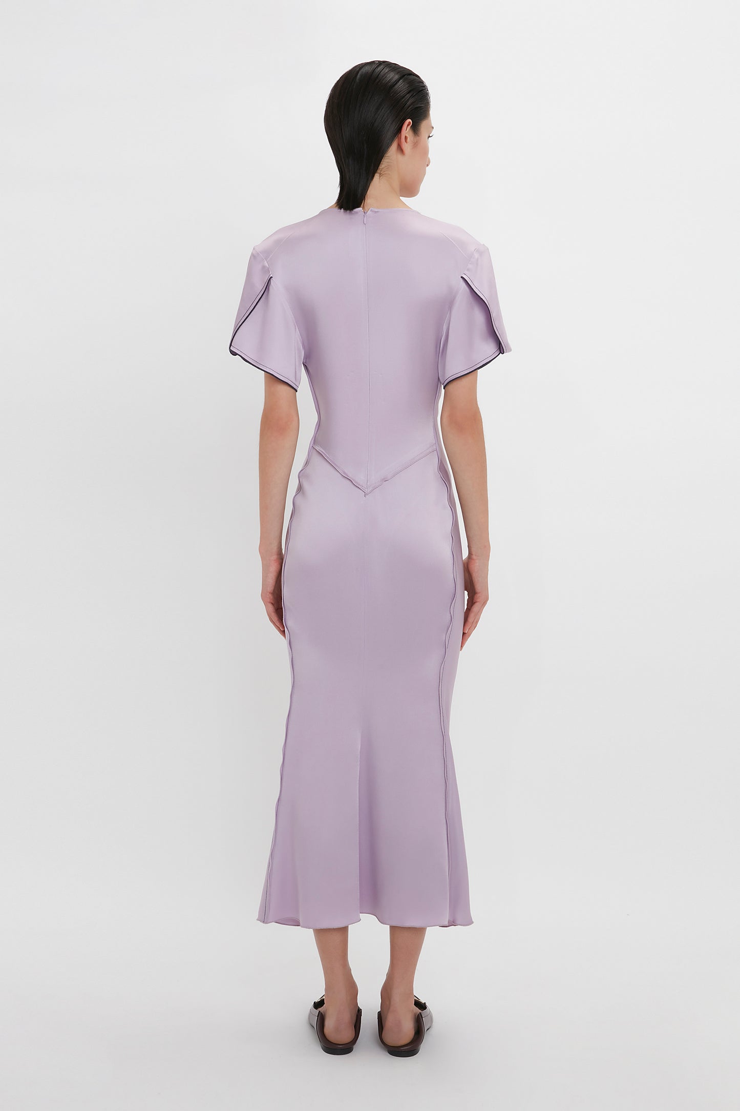 A person is standing facing away from the camera, wearing a light purple, form-fitting Gathered V-Neck Midi Dress In Petunia with short sleeves and black shoes. The elegant dress channels Victoria Beckham's timeless style against a plain white background.