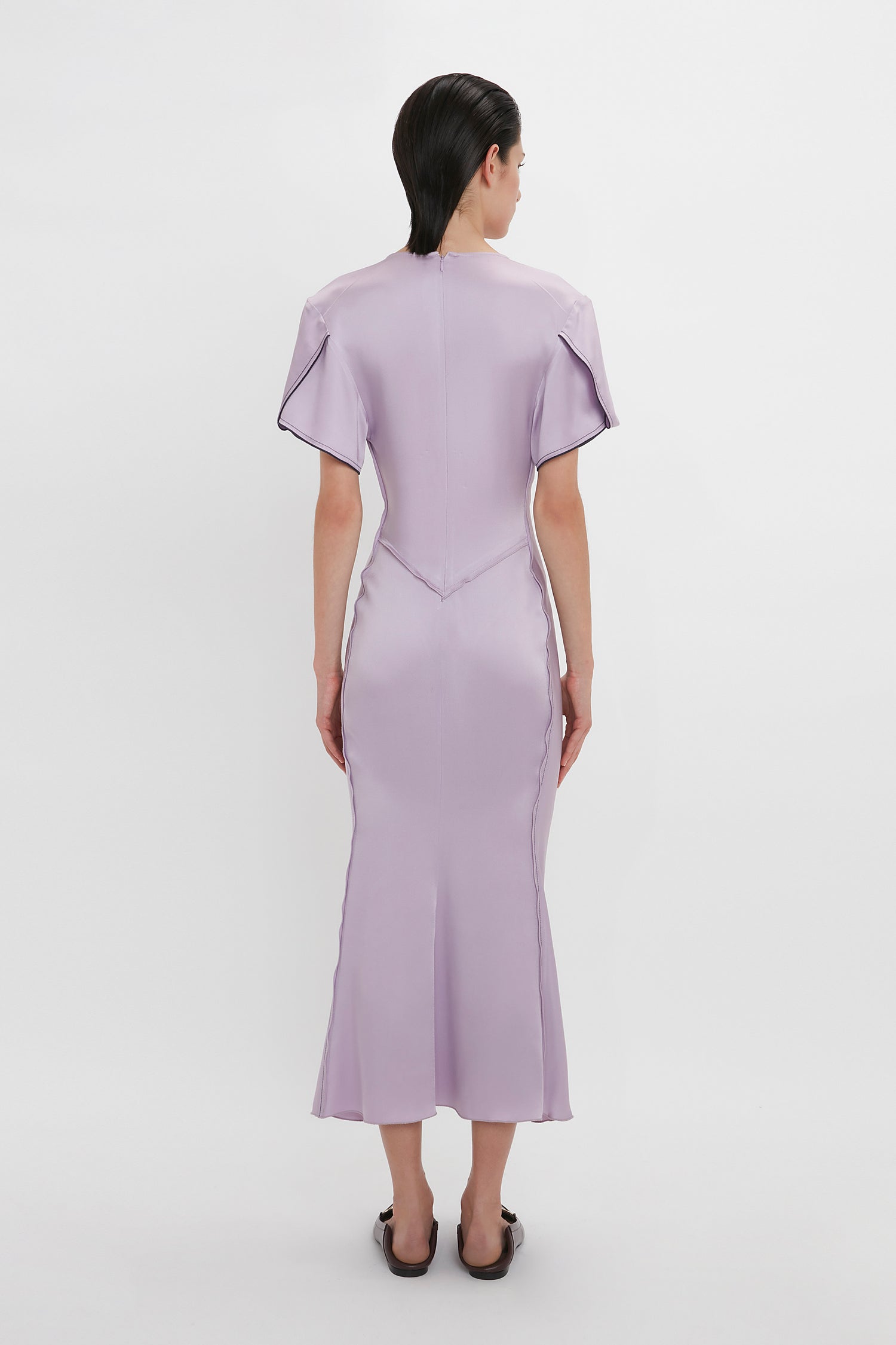 A person is standing facing away from the camera, wearing a light purple, form-fitting Gathered V-Neck Midi Dress In Petunia with short sleeves and black shoes. The elegant dress channels Victoria Beckham's timeless style against a plain white background.