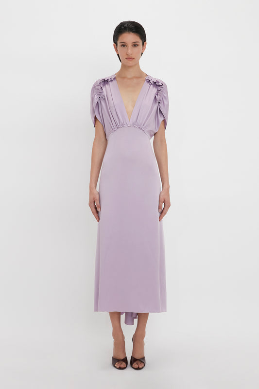 A person wearing a light purple, Victoria Beckham V-Neck Ruffle Midi Dress In Petunia stands against a plain white background. They have short dark hair and are sporting dark open-toe shoes.