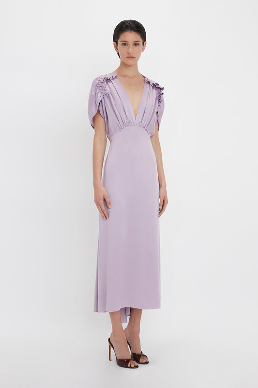 A person is wearing the Victoria Beckham V-Neck Ruffle Midi Dress In Petunia with short, draped sleeves and a deep V-neckline. Reminiscent of the Victoria Beckham brand’s sophisticated touch, they are standing against a plain, white background.