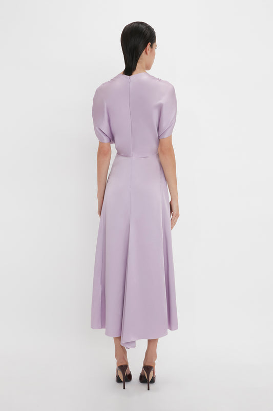 A woman with dark hair wearing an elegant light purple, short-sleeved dress and black heels is standing with her back to the camera on a plain white background, showcasing the timeless style of the V-Neck Ruffle Midi Dress In Petunia by Victoria Beckham.