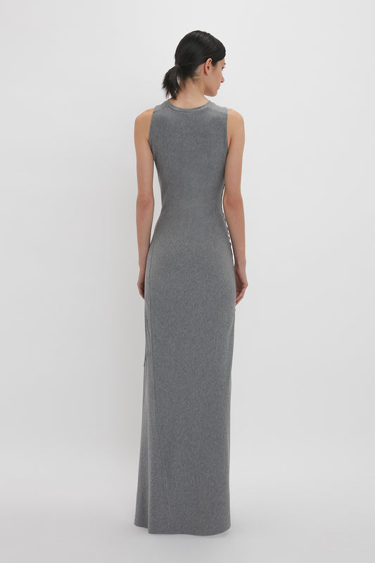 A woman viewed from behind, wearing a long gray sleeveless Victoria Beckham Frame Detailed Maxi Dress In Titanium with a high neckline, standing against a white background.