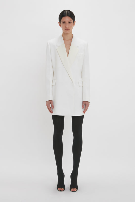 A woman in an Exclusive Fold Shoulder Detail Dress In Ivory by Victoria Beckham paired with black tights and heels stands against a plain white background.