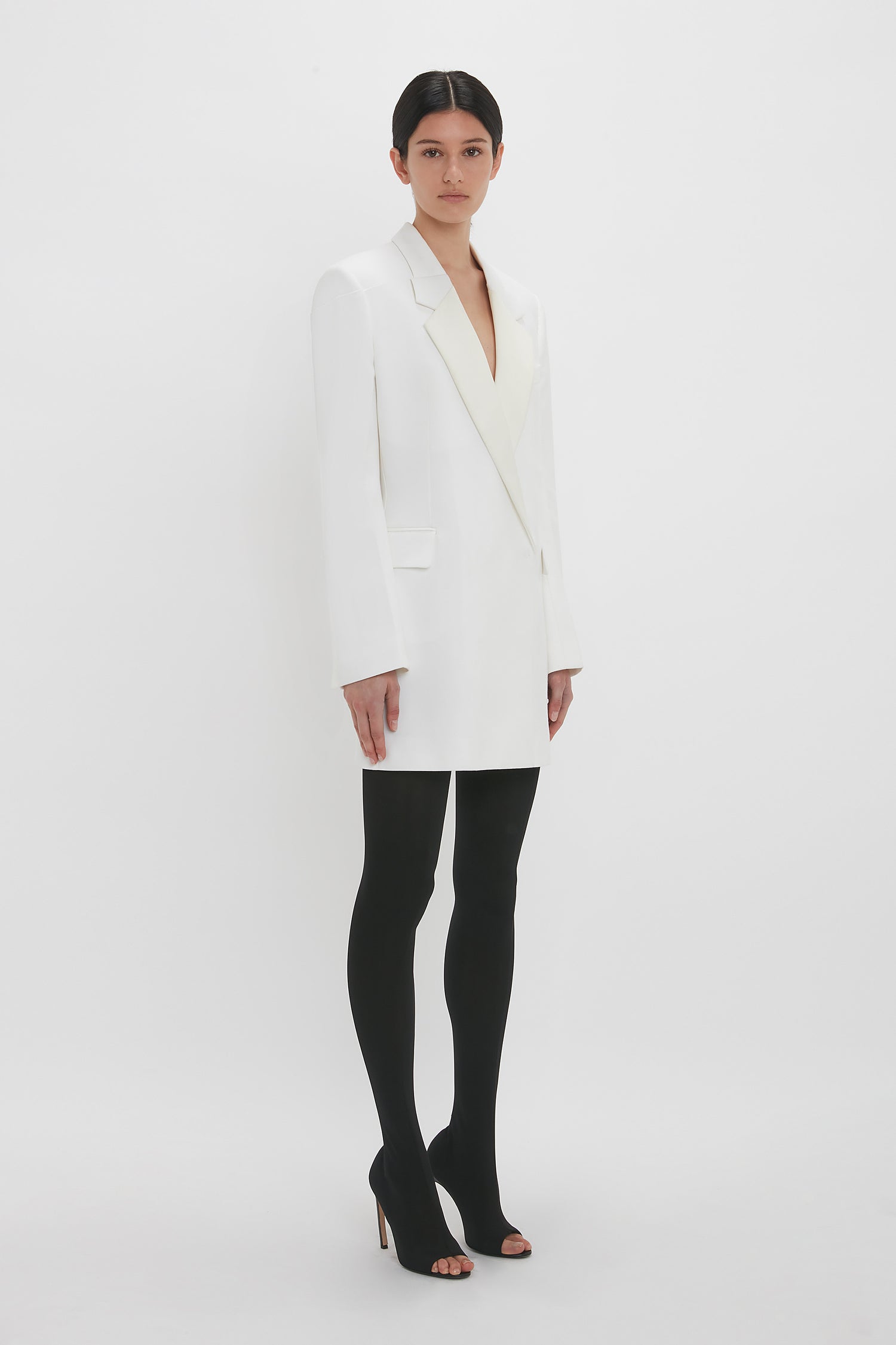 A woman stands against a white background, wearing a Victoria Beckham Exclusive Fold Shoulder Detail Dress In Ivory, black tights, and black high heels, looking directly at the camera.