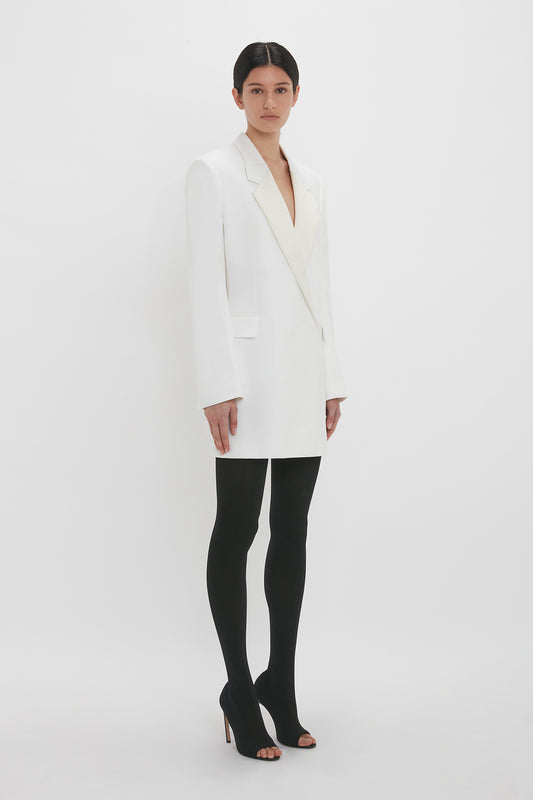 A woman stands against a white background, wearing a Victoria Beckham Exclusive Fold Shoulder Detail Dress In Ivory, black tights, and black high heels, looking directly at the camera.