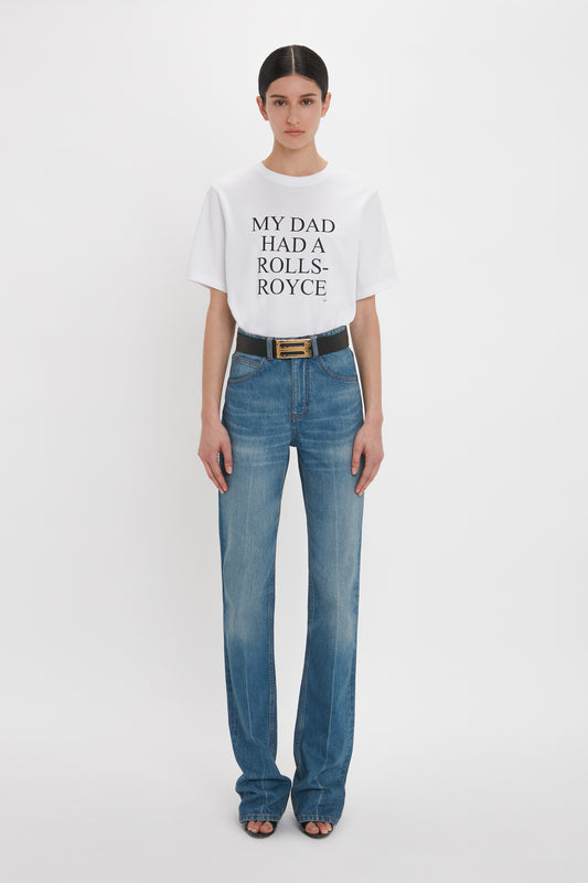 A woman in a white t-shirt with text "my dad had a Rolls-Royce" and blue distressed high-waisted Victoria Beckham Julia Jean In Broken Vintage Wash, standing against a plain background.