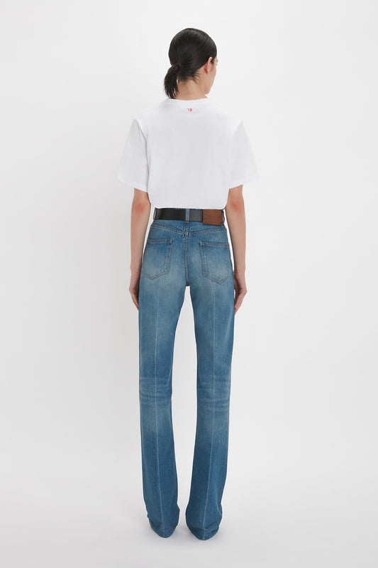 A person seen from behind, wearing a white t-shirt and Julia Jean In Broken Vintage Wash high waist jeans with a black belt, standing against a plain white background by Victoria Beckham.