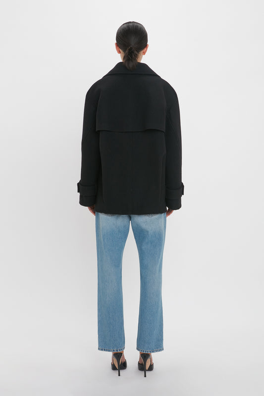Person standing facing away, wearing an Oversized Pea Coat In Black by Victoria Beckham, light blue jeans, and black high-heeled shoes against a plain white background.