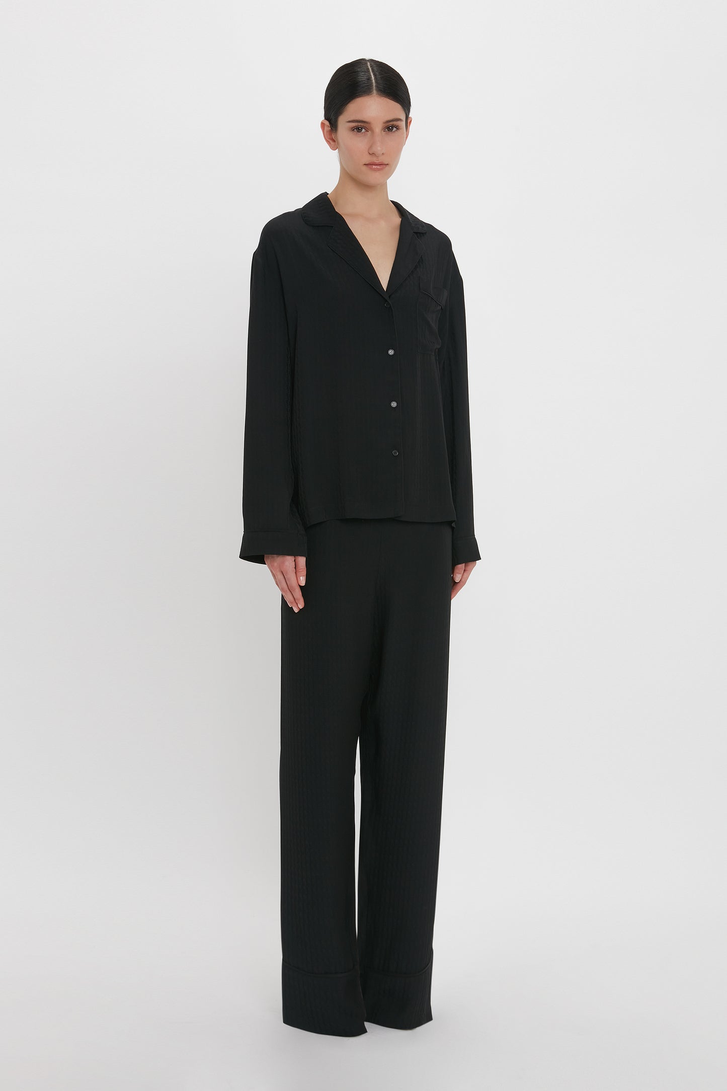 Person standing against a plain white background wearing a loose-fitting VB Monogram Pyjama Sleep Set In Black by Victoria Beckham, accessorized with a matching tote and eye mask.