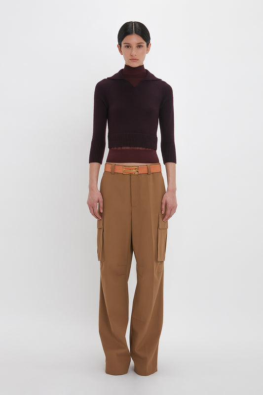 Person stands against a white background, wearing a dark maroon, collarless top with elbow-length sleeves, Victoria Beckham Relaxed Cargo Trouser In Tobacco, and a tan belt with an elevated detailing gold and brown buckle.