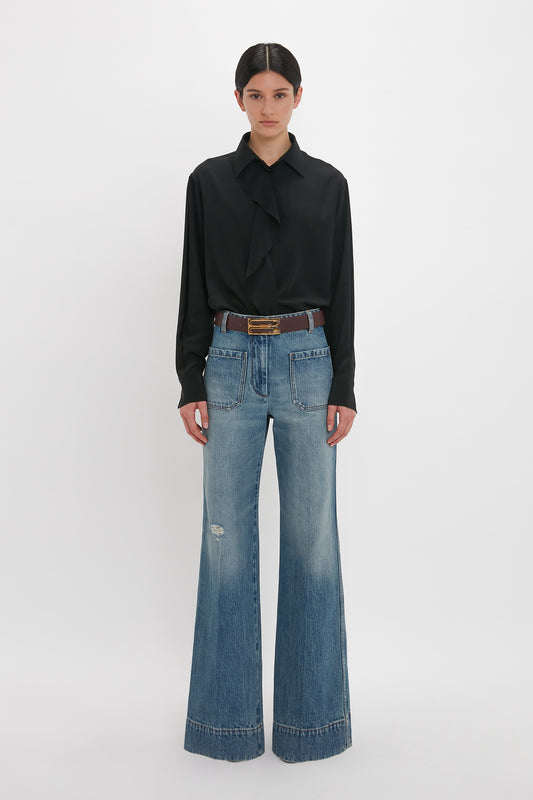 A person stands against a white background, embodying contemporary timeless fashion with the Victoria Beckham Asymmetric Ruffle Blouse In Black and light blue wide-legged jeans cinched with a belt.
