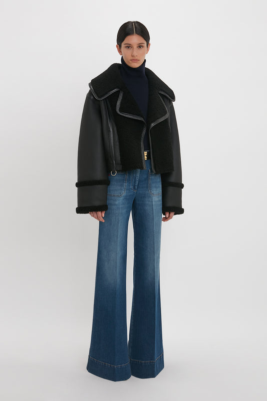 A woman stands wearing a luxurious Victoria Beckham shearling aviator jacket in black, blue turtleneck, and flared jeans against a white background.