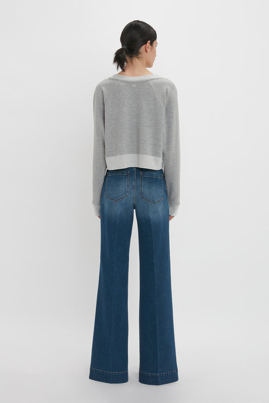 A woman stands facing away from the camera, wearing a Victoria Beckham cropped sweatshirt in grey marl and flared blue jeans, against a plain white background.