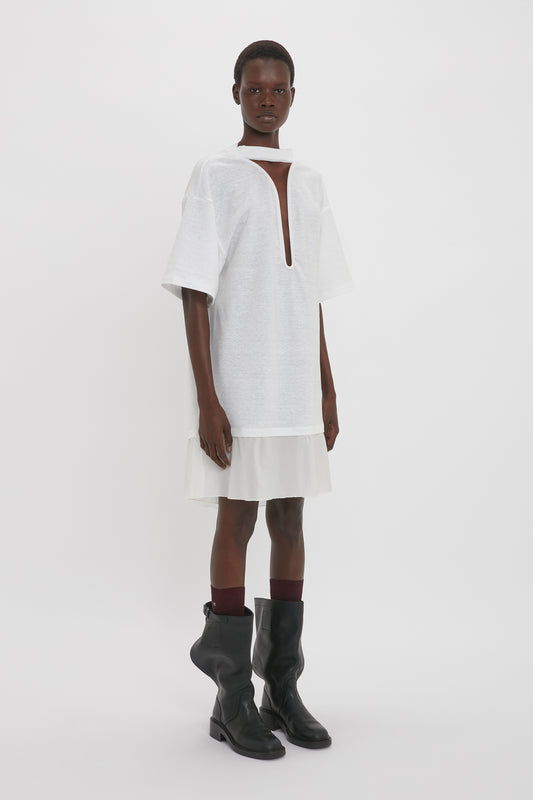 A black woman models a Victoria Beckham Frame Cut-Out T-Shirt Dress in White with a keyhole neckline, paired with dark knee-high boots against a plain background.