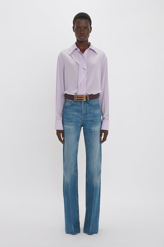 A tall model standing against a white background, wearing a Victoria Beckham Asymmetric Ruffle Blouse in Petunia color tucked into high-waisted blue jeans with a brown belt.