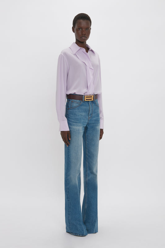 A person standing against a white background wearing a Victoria Beckham Asymmetric Ruffle Blouse in Petunia, blue jeans, and a yellow belt.