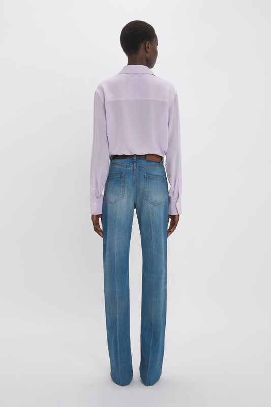 A person stands with their back to the camera, wearing a Victoria Beckham Asymmetric Ruffle Blouse in Petunia and classic blue jeans, against a plain white background.