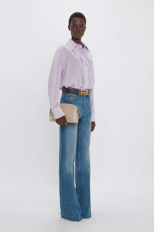 A black woman stands wearing a Victoria Beckham asymmetric ruffle blouse in petunia, blue jeans, and a tan belt, holding a beige shoulder bag, against a white background.
