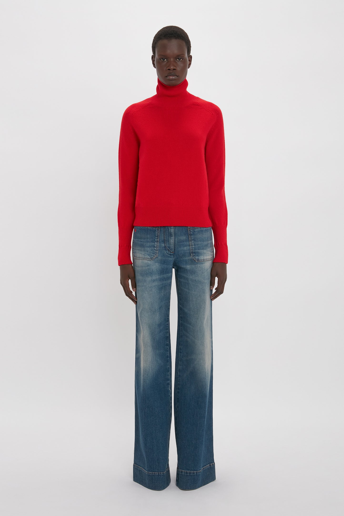 A black man wearing a red turtleneck sweater and Victoria Beckham's Alina Jean In Heavy Vintage Indigo Wash stands against a white background.