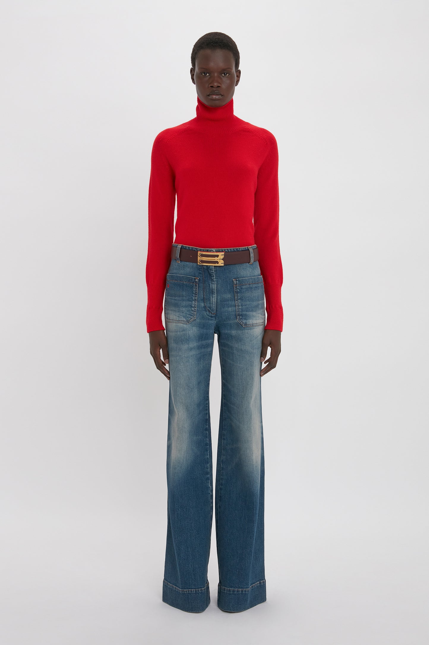 A black woman stands against a white background, wearing a red turtleneck sweater, Victoria Beckham's Alina Jean in Heavy Vintage Indigo Wash, and a brown belt.
