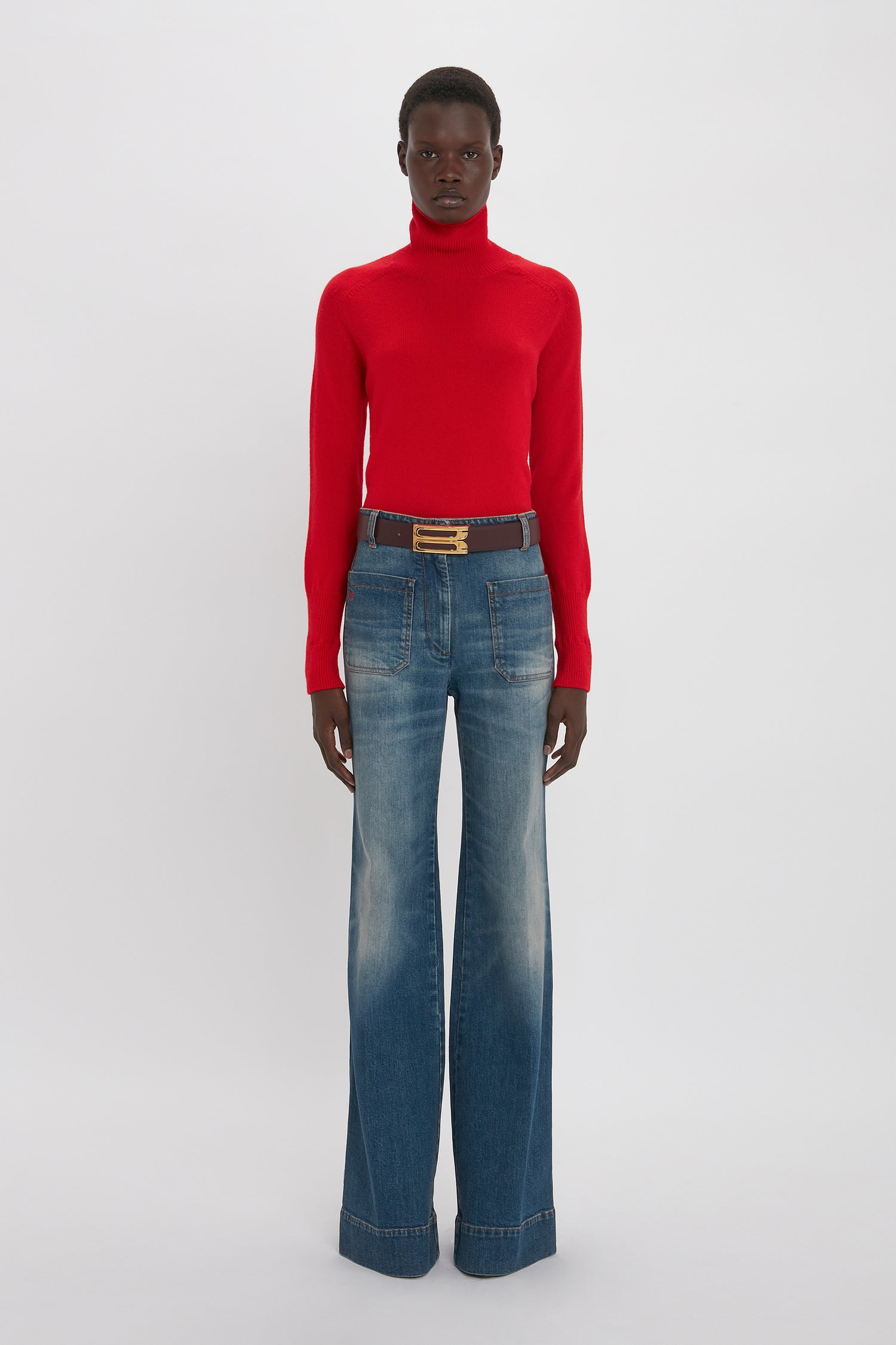 A black woman stands against a white background, wearing a red turtleneck sweater, Victoria Beckham's Alina Jean in Heavy Vintage Indigo Wash, and a brown belt.