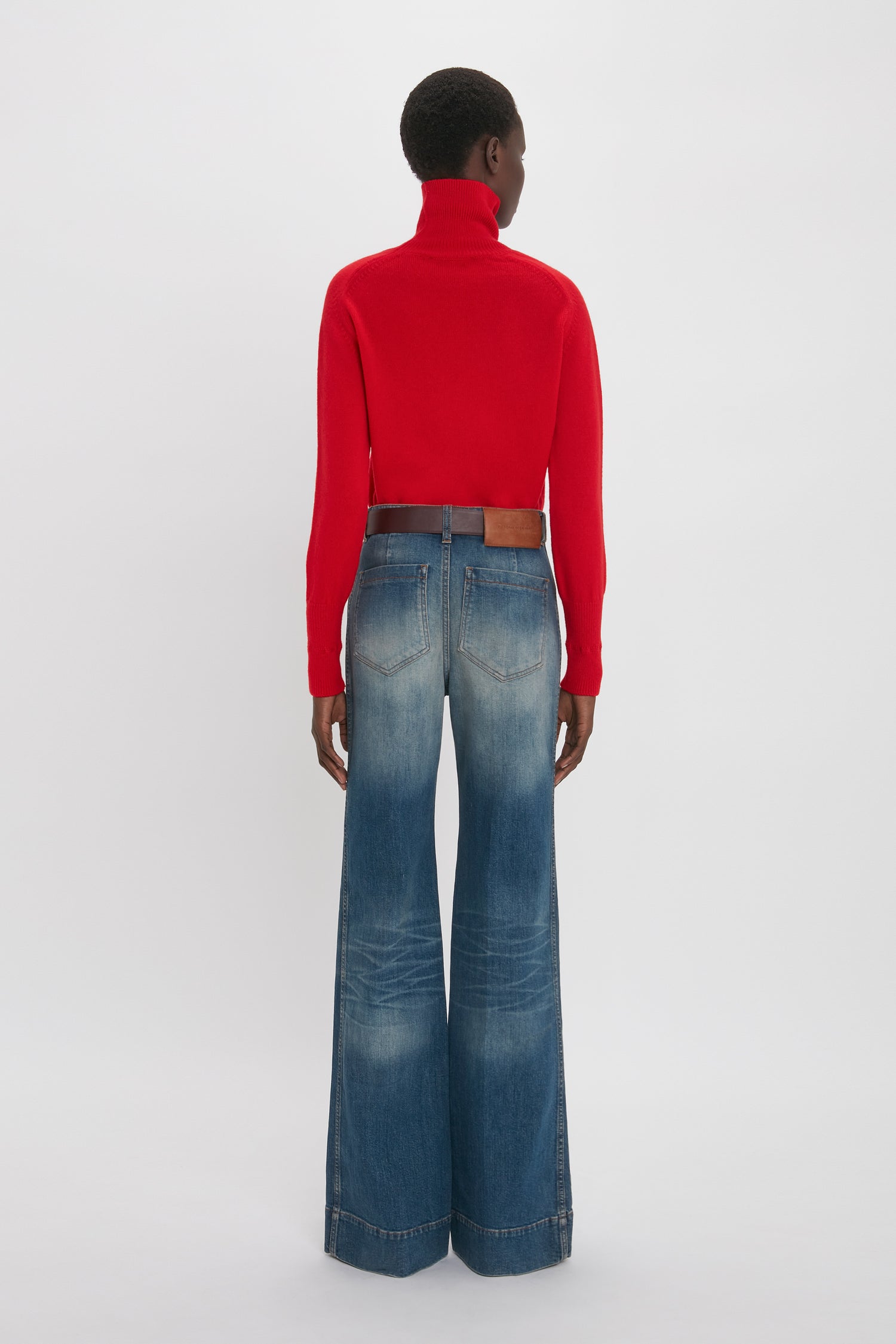 A woman stands with her back to the camera, wearing a red turtleneck and blue Victoria Beckham Alina Jean in Heavy Vintage Indigo Wash, against a plain white background.