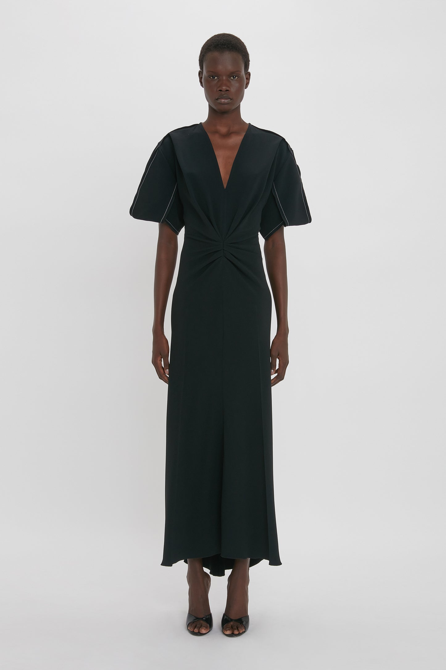 A person stands against a white background wearing a Gathered V-Neck Midi Dress in Black from Victoria Beckham with short, wide sleeves, paired with black open-toe shoes. Made from figure-flattering stretch fabric, the black dress exudes a contemporary edge while their hands are relaxed by their sides.