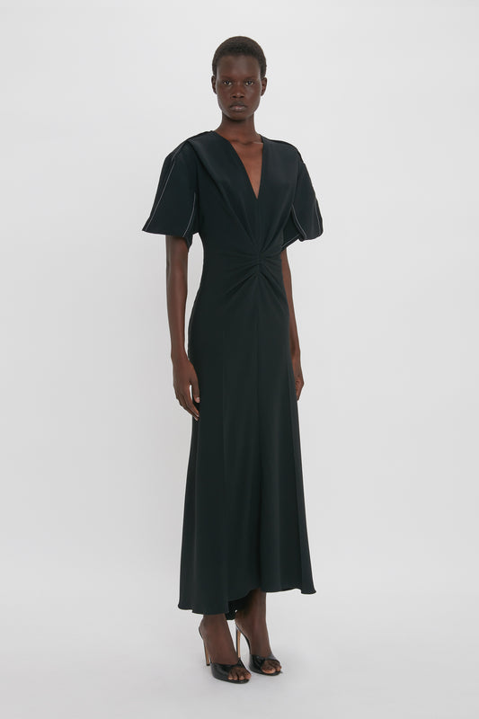 A person wearing a black, short-sleeve, Gathered V-Neck Midi Dress in Black by Victoria Beckham stands facing the camera in a white studio background, exuding a contemporary edge.