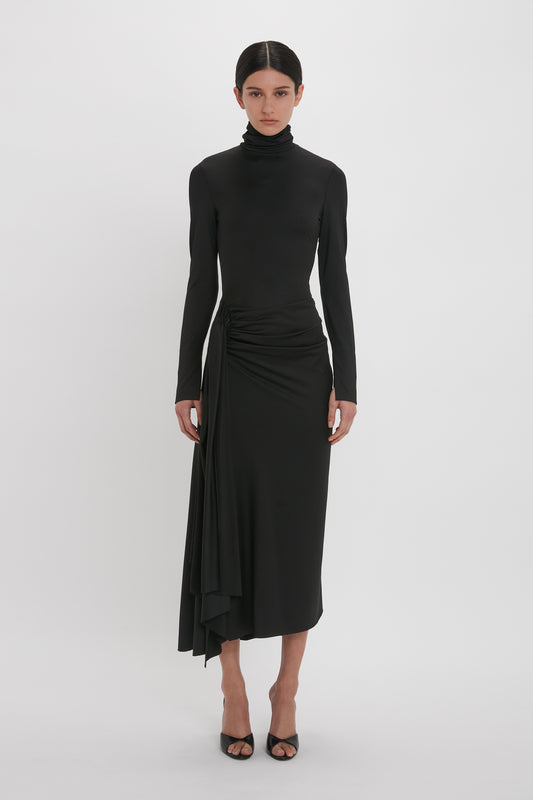 A woman in a Victoria Beckham High Neck Asymmetric Draped Dress In Black with an asymmetric hem and open-toe heels stands against a plain white background.