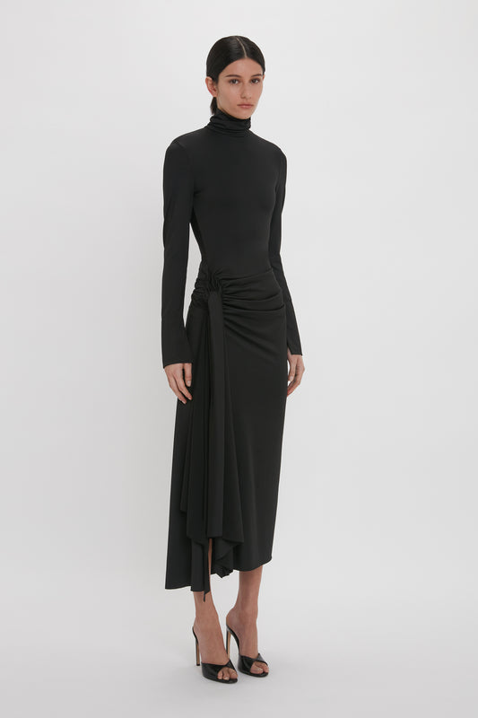 A woman in a sleek Victoria Beckham high neck asymmetric draped dress in black with an asymmetric hem and high slit, paired with black heels, posing against a white background.