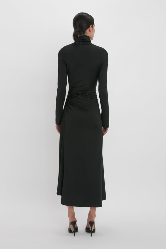 A woman in a black Victoria Beckham High Neck Asymmetric Draped Dress, with a cinched waist and asymmetric hem, seen from behind against a plain white background.