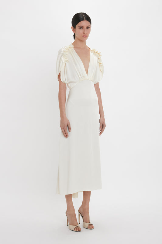 A woman stands against a white background wearing a Victoria Beckham Exclusive V-Neck Ruffle Midi Dress In Ivory with gathered short sleeves, paired with open-toe high heels. She has dark hair pulled back.
