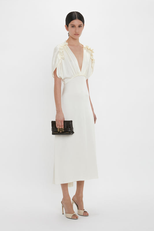 A woman with dark hair in a low bun is wearing a white Exclusive V-Neck Ruffle Midi Dress In Ivory by Victoria Beckham. The mid-length dress features ruffle details on the shoulders. She is holding a black clutch and wearing open-toed heels.