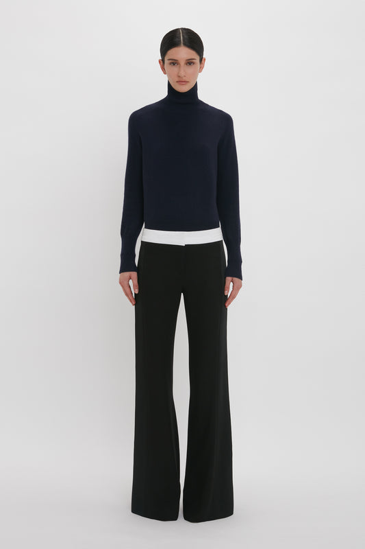 A person stands against a plain white background, wearing a dark navy turtleneck sweater, white undershirt, and the Side Panel Trouser In Black by Victoria Beckham with a wide-leg design. They have dark hair tied back.