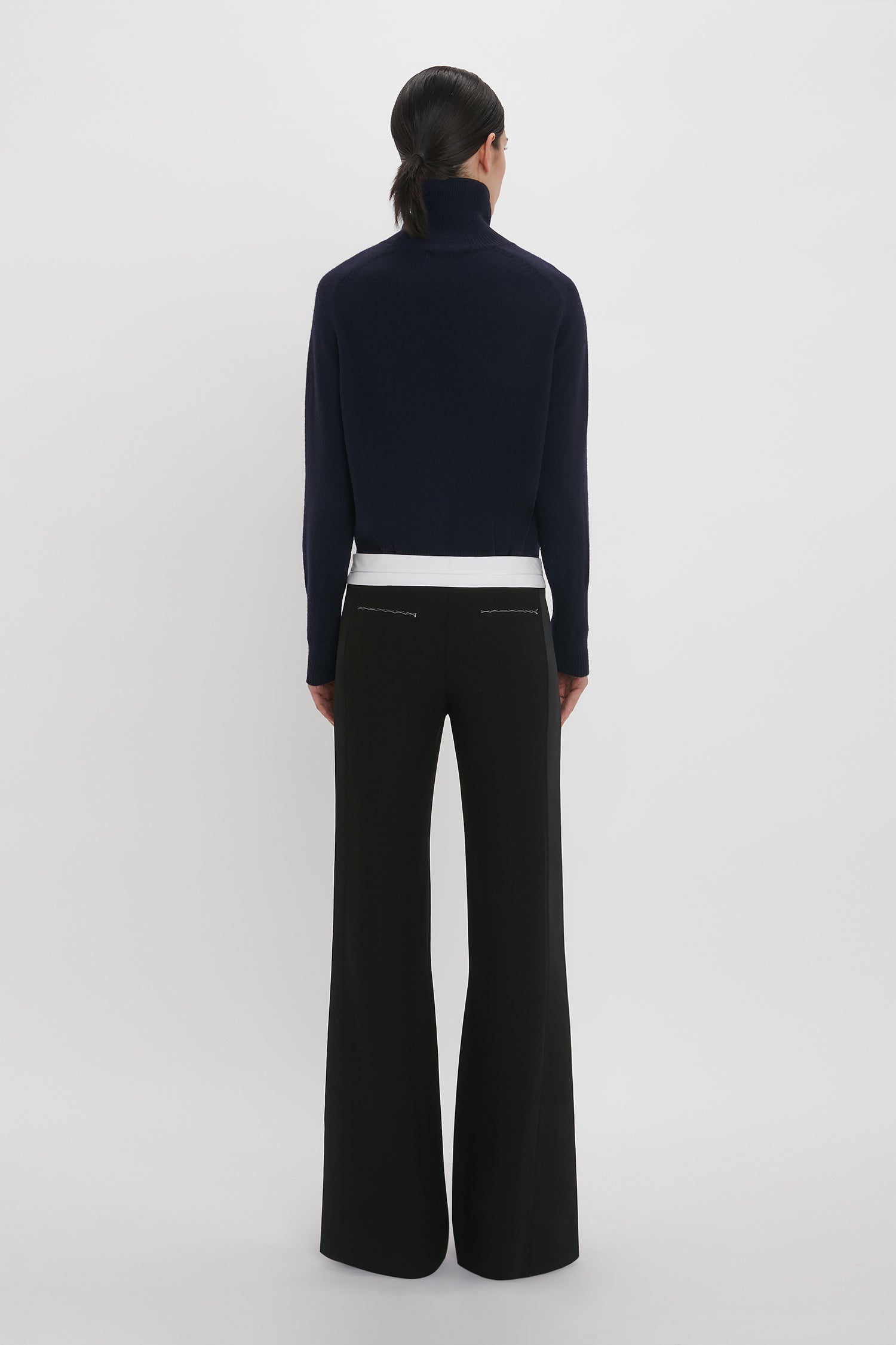 A person with dark hair is standing with their back to the camera, wearing a black turtleneck sweater, Victoria Beckham Side Panel Trouser In Black with a wide-leg design, and a white top underneath.