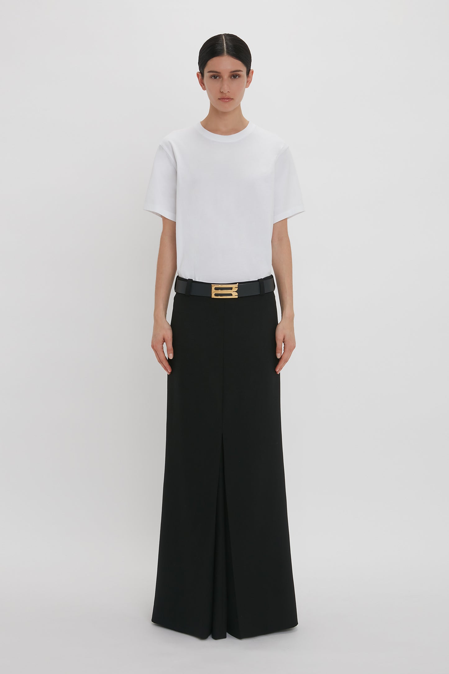 A woman standing in a white studio, wearing a Victoria Beckham Floor-Length Box Pleat Skirt in Black and high-waisted black trousers with a broad belt with gold buckle.