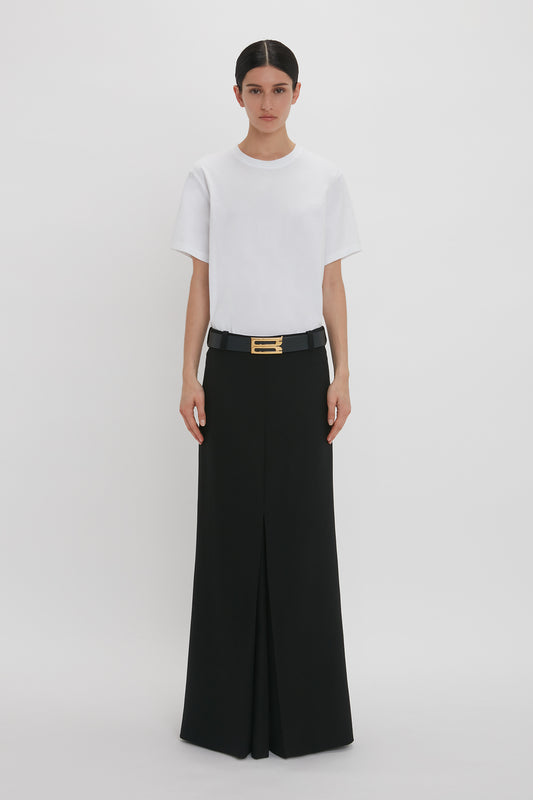 A woman standing in a white studio, wearing a Victoria Beckham Floor-Length Box Pleat Skirt in Black and high-waisted black trousers with a broad belt with gold buckle.