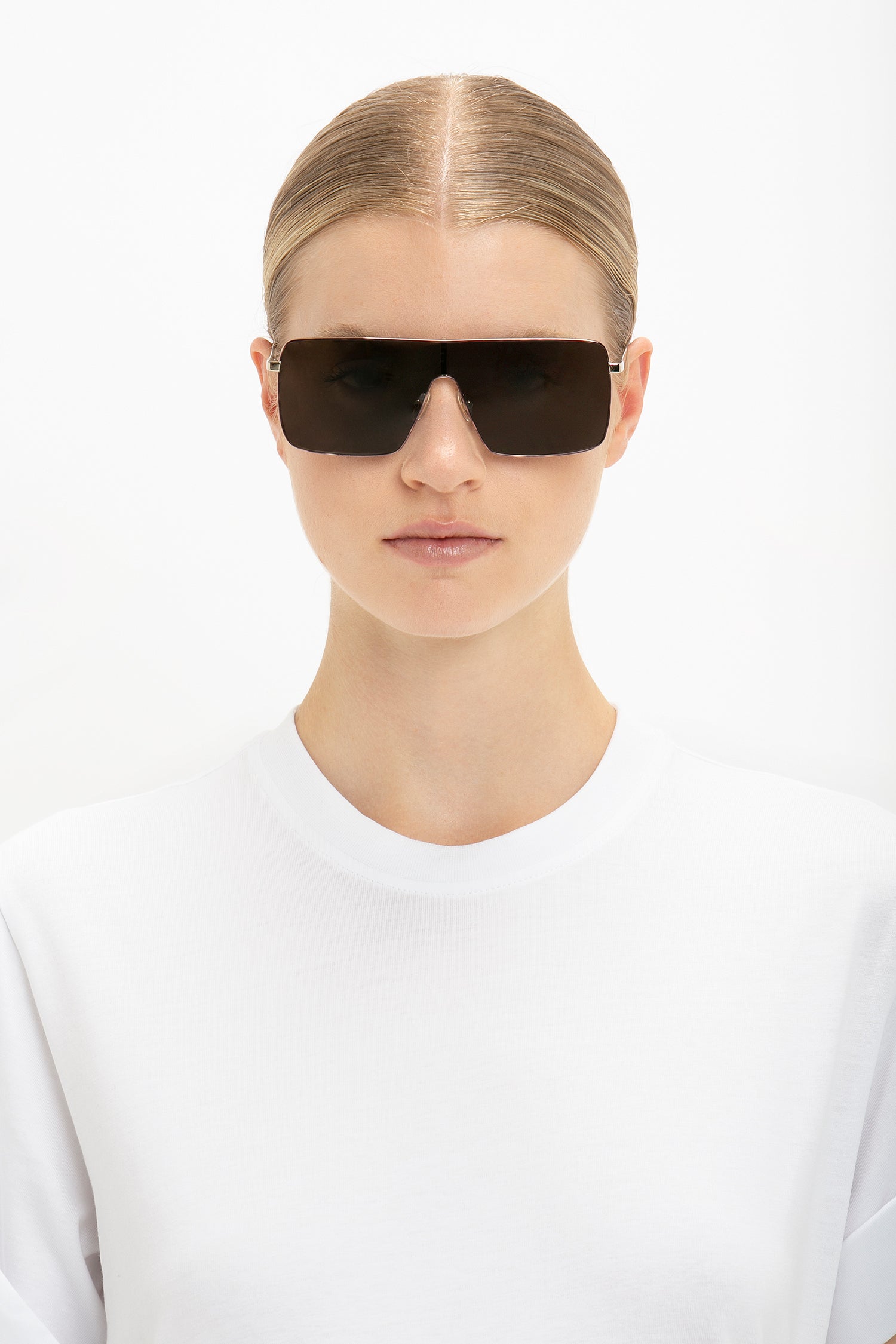 Person with blond hair wearing Victoria Beckham's Rectangular Visor In Gold-Smoke sunglasses and a white top against a plain background.