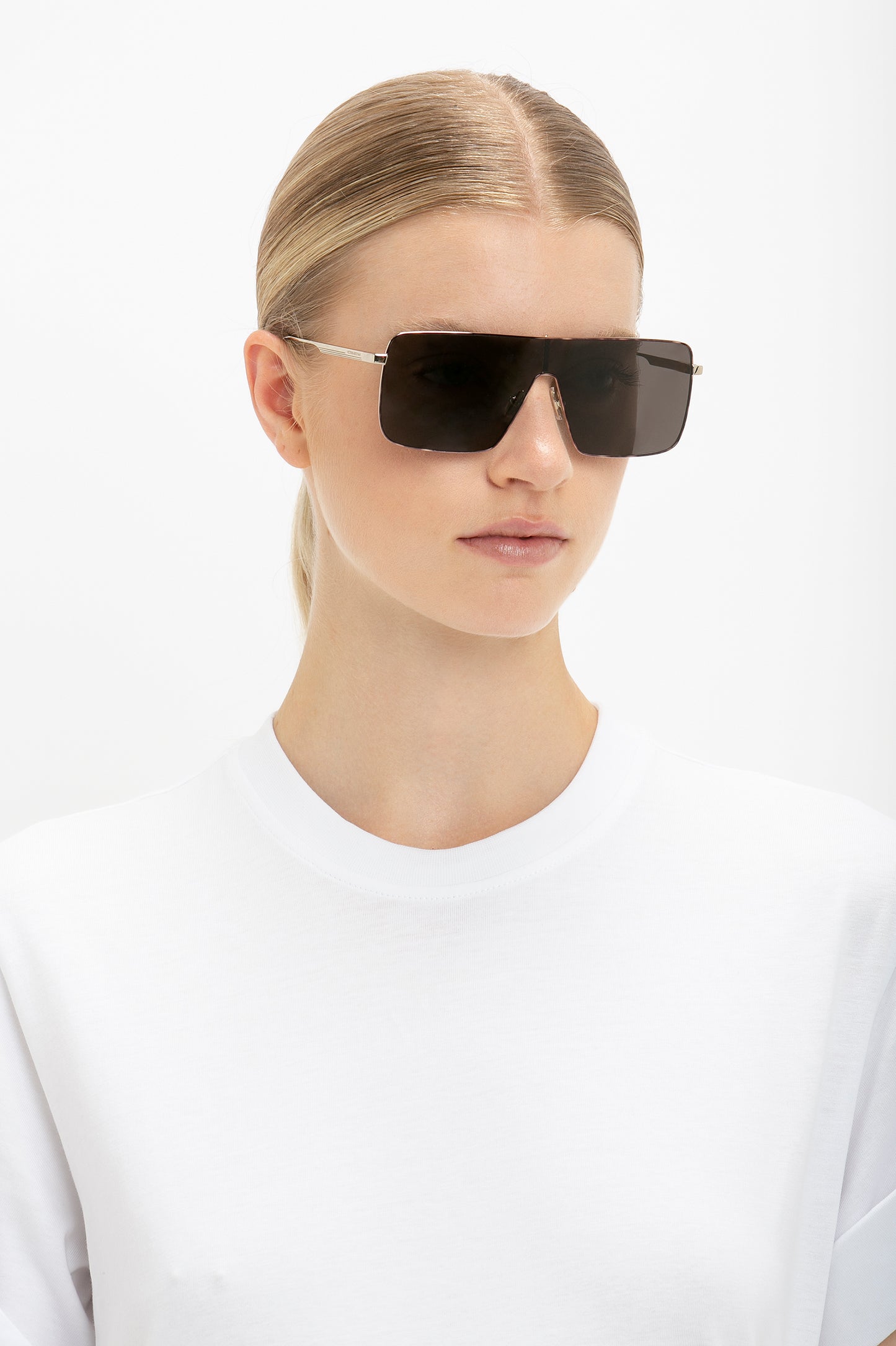 Person with blonde hair wearing a white shirt and large Rectangular Visor In Gold-Smoke by Victoria Beckham, looking forward against a plain white background.