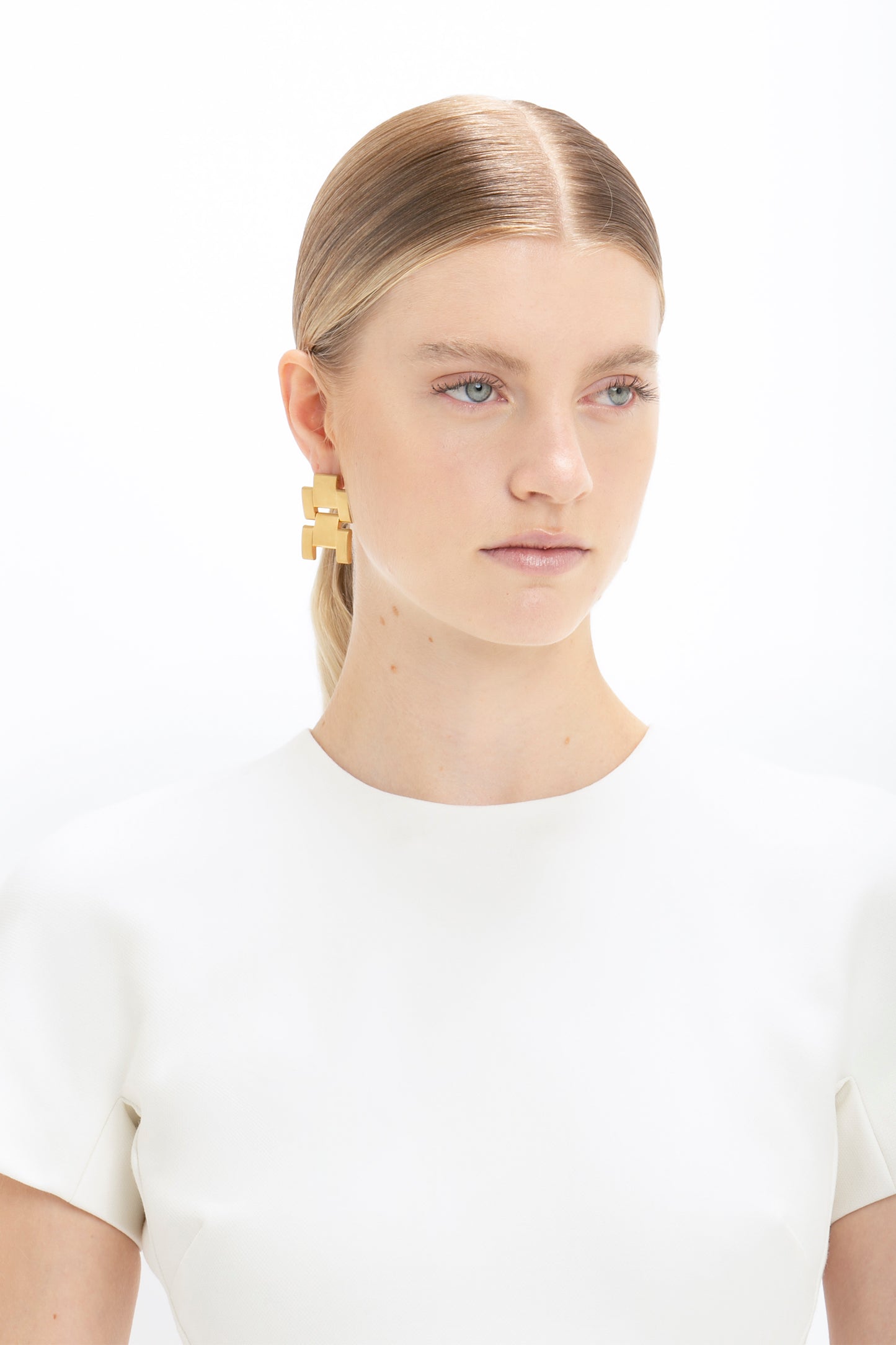 A woman with slicked-back hair wearing a Victoria Beckham fitted T-shirt dress in ivory and a large gold earring shaped like a puzzle piece, against a white background.